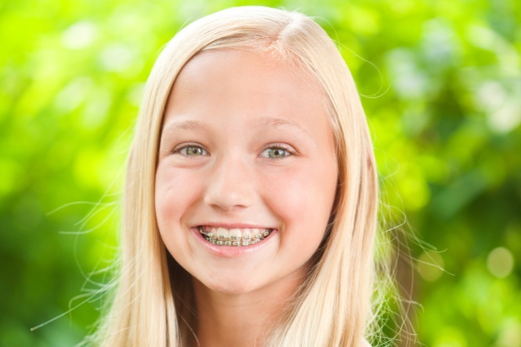 Blonde girl wearing braces smiles against bright green foliage