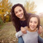 Two smiling sisters with braces riding piggyback in a park.