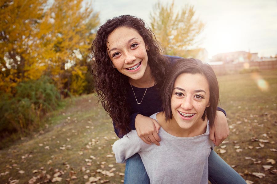 Two smiling sisters with braces riding piggyback in a park.