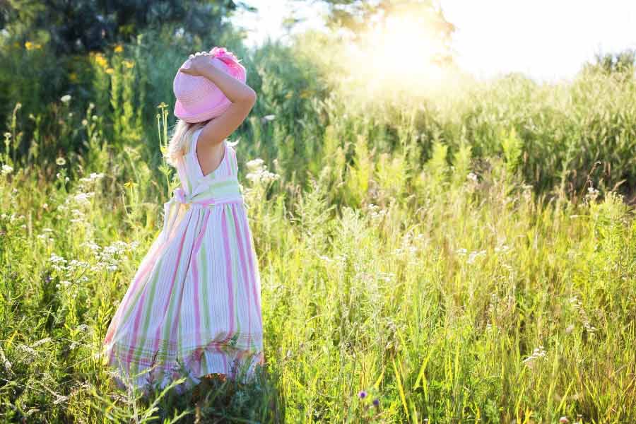 Young girl in a meadow with a long dress and floppy pink hat facing away.