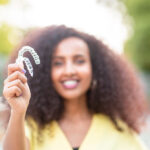 A Black woman with curly hair in a yellow shirt smiles while holding up her 2 Invisalign clear aligners outside in Littleton, CO