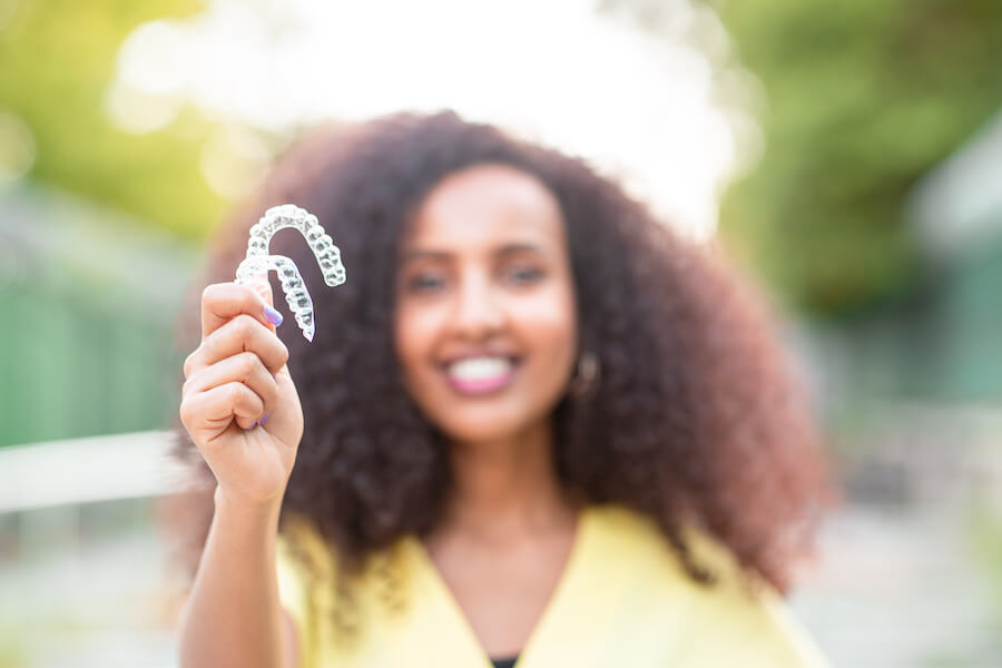 A Black woman with curly hair in a yellow shirt smiles while holding up her 2 Invisalign clear aligners outside in Littleton, CO