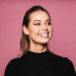 Brunette woman in a black sweater smiles with straight teeth against a pink wall