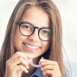 Young teen with black rimmed glasses smiles while holding her Invisalign clear aligners