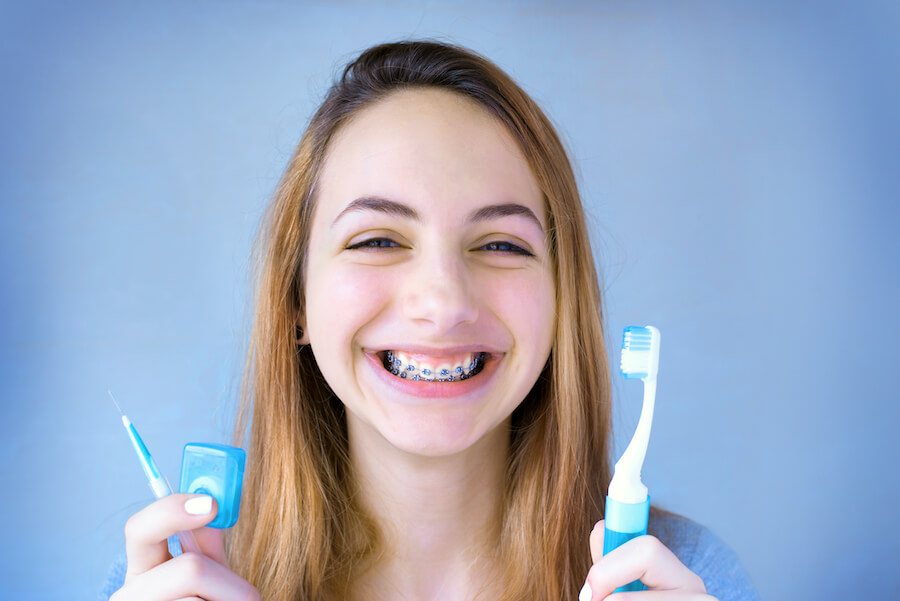 Young woman smiles with braces as she holds up her toothbrush, floss, and interdental brush to clean her teeth